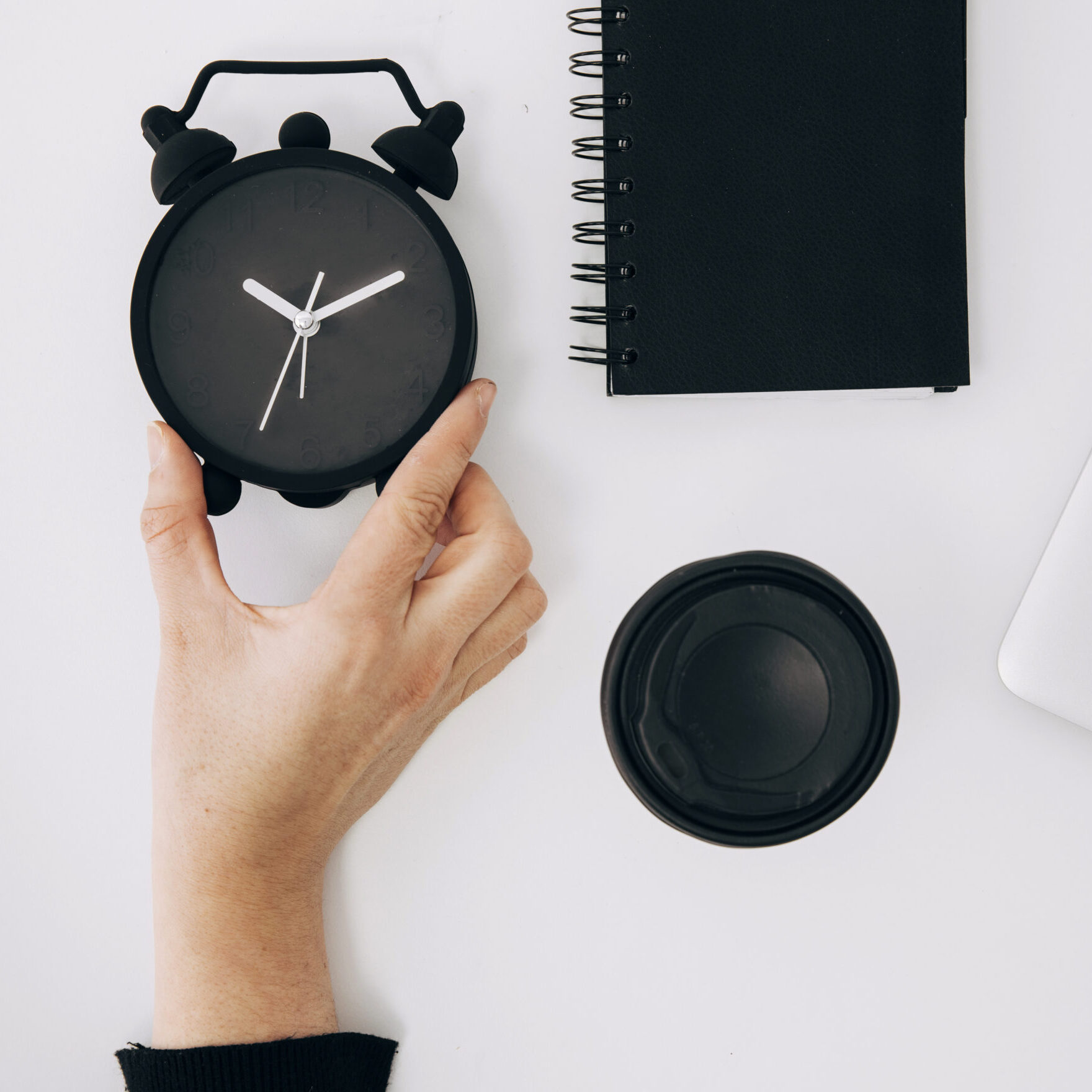 close up person s hand holding black alarm clock spiral notepad laptop takeaway coffee cup white desk